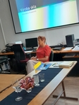 Přednášející (členka SONS)  sedí na židli při výkladu - v popředí ukázka různých druhů skla